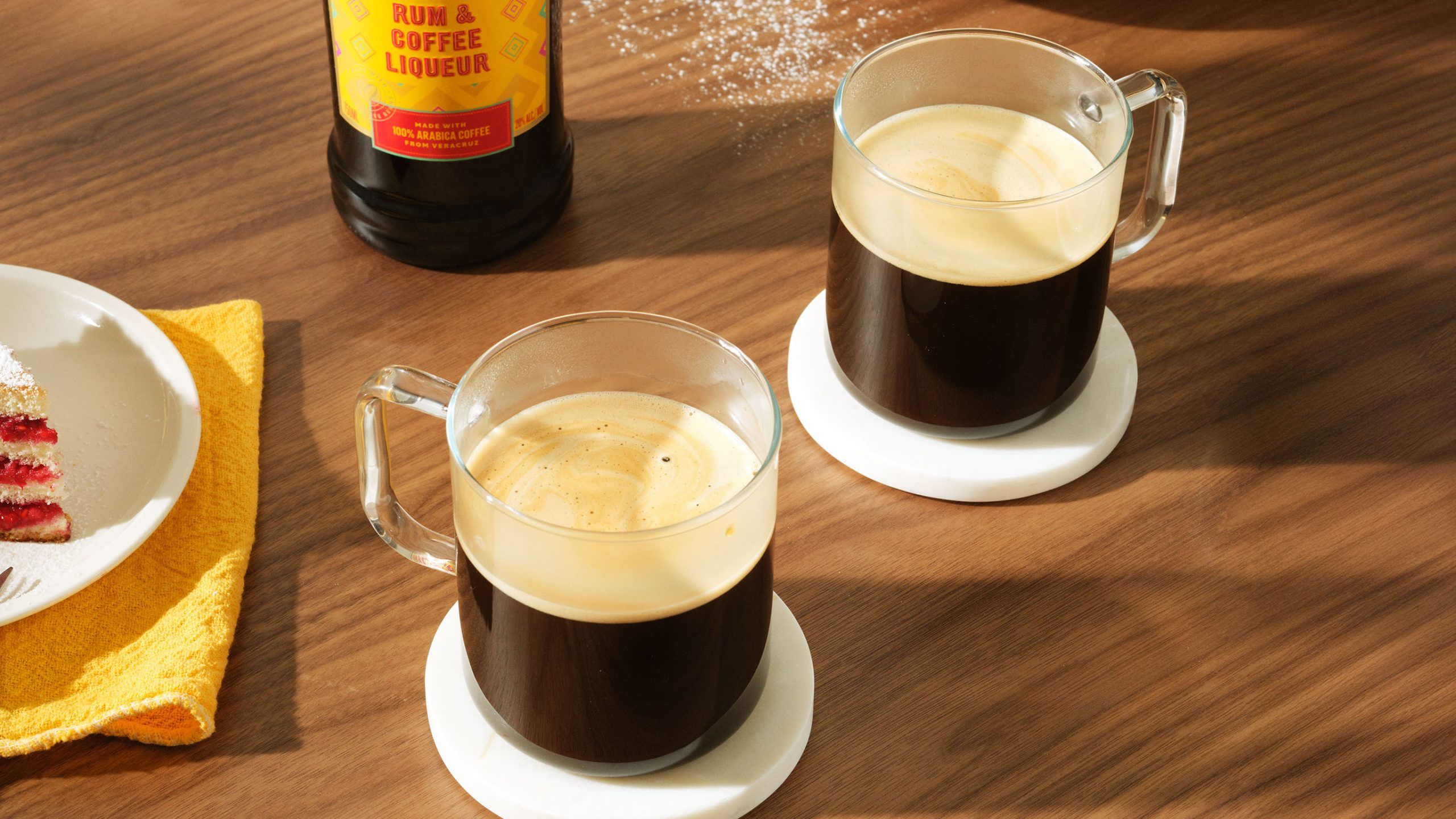 Two kahlua coffee drinks on a dining table, with a kahlua bottle and cake slices