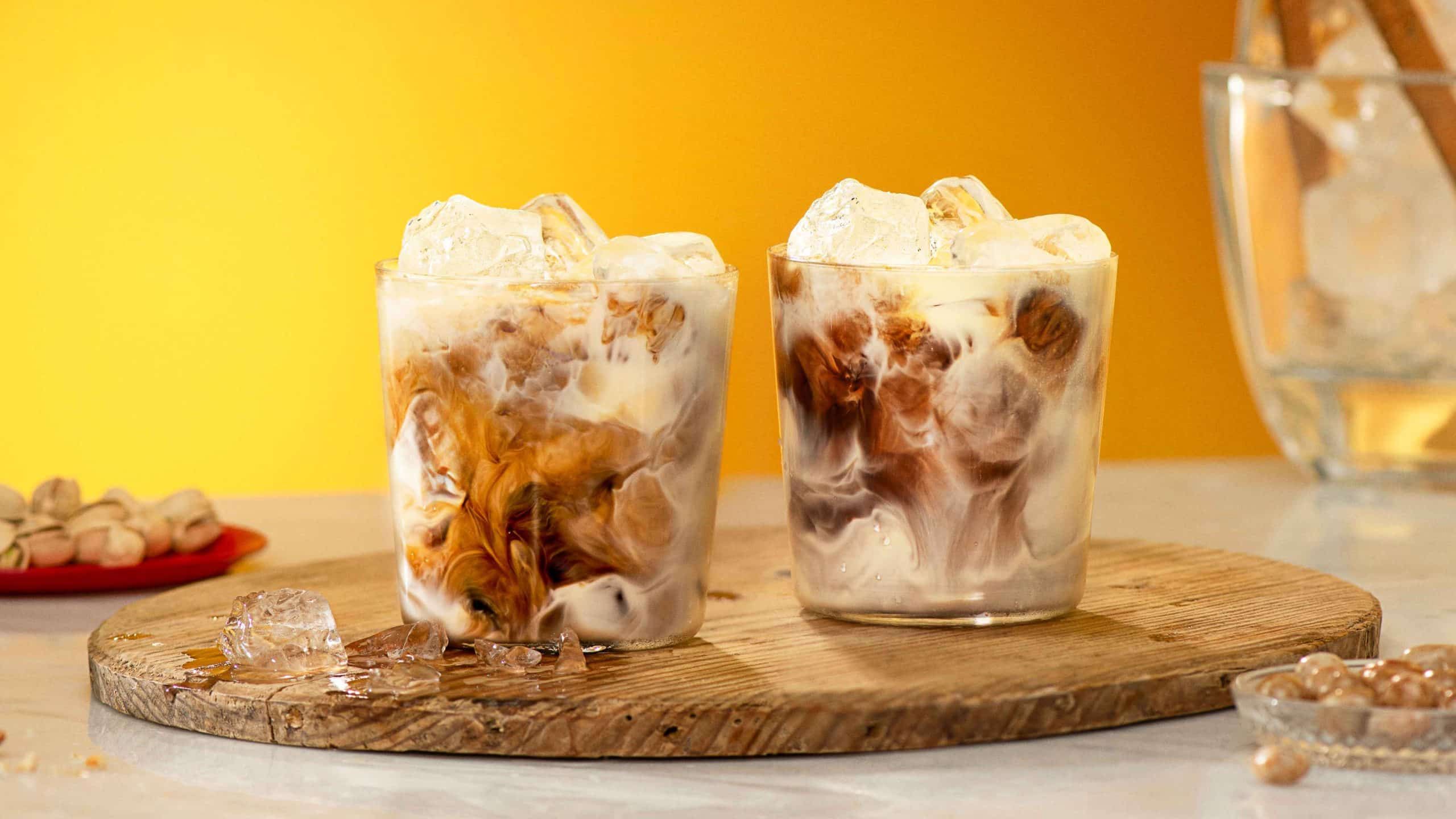 Two drinks with Kahlúa and milk garnished with ice cubes in front of a yellow background