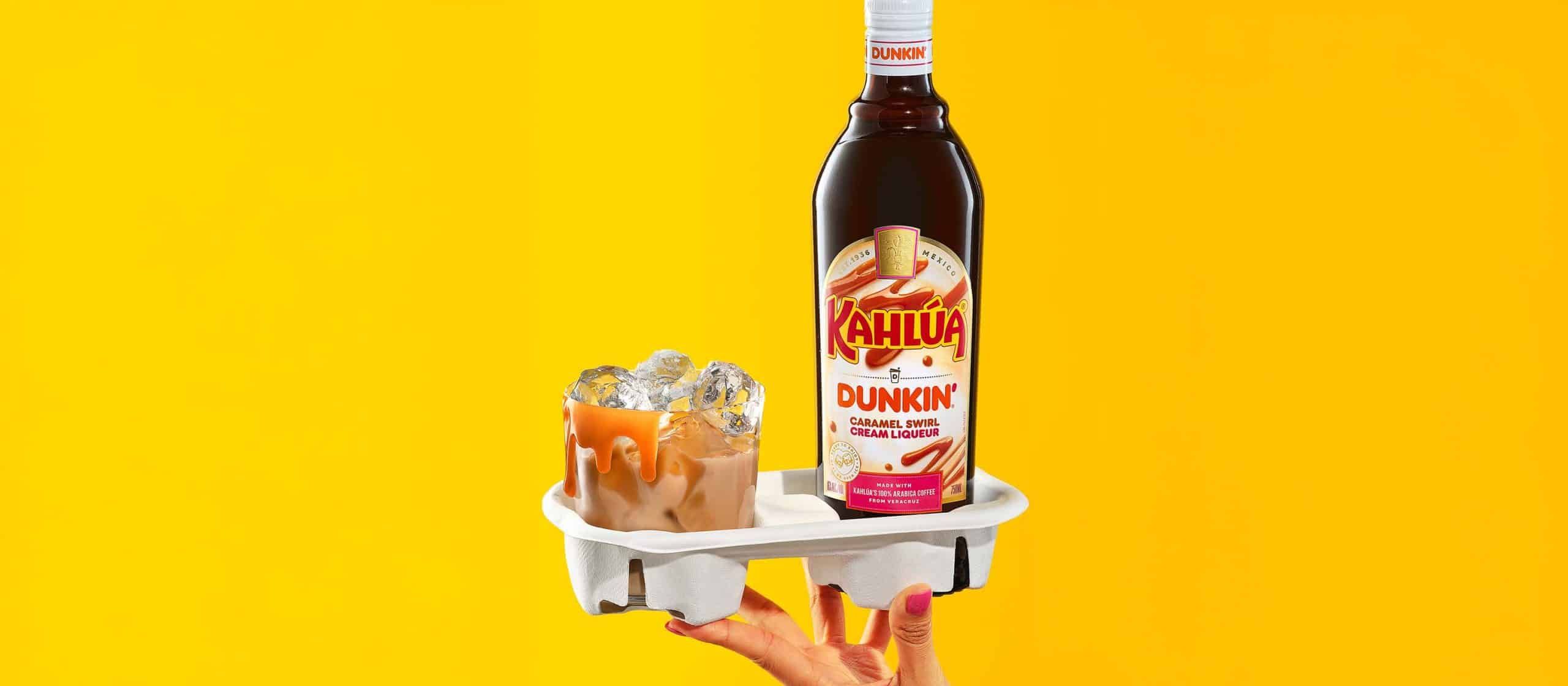 2 items being held in a cardboard coffee cup holder against a bright yellow background- First a brown cocktail in a rocks glass with ice. Second, a bottle of kahlua dunkin caramel swirl
