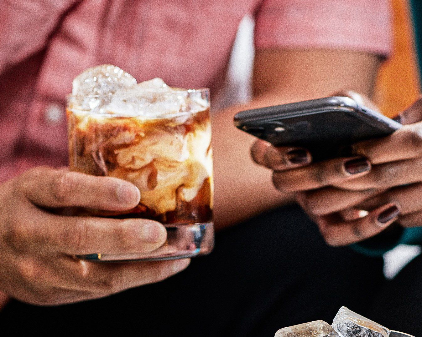 Two pair of hands in frame. One person holding a white russian, another scrolling on a black phone